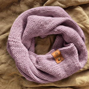 Könnte beinhalten: Ein lavendelfarbener Infinity-Schal aus grobem Strickgarn. Der Schal hat ein kleines, rechteckiges, hellbraunes Lederetikett, das mit einer silberfarbenen Metallniete befestigt ist. Das Etikett zeigt eine Kaffeetasse und den Text "Dandelion Knits".