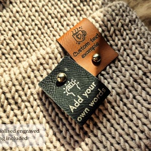 Op de afbeelding: Twee rechthoekige kunstlederen labels bevestigd op beige gebreide stof. E&eacute;n label is bruin met een koffiekop-icoon en de tekst "Custom text example". Het andere label is zwart met een hand-icoon en de tekst "Add your own words". Beide hebben een zilveren klinknagel.