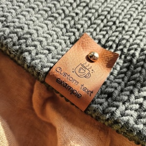 Op de afbeelding: Een donkergrijze gebreide stof met een beige leren label bevestigd met een metalen klinknagel. Het label bevat een afbeelding van een koffiemok met een hartje en de tekst "Custom text example" in zwarte inkt op het leer gedrukt.