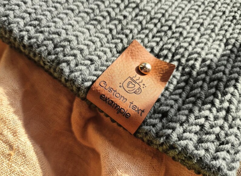 Op de afbeelding: Een donkergrijze gebreide stof met een beige leren label bevestigd met een metalen klinknagel. Het label bevat een afbeelding van een koffiemok met een hartje en de tekst "Custom text example" in zwarte inkt op het leer gedrukt.