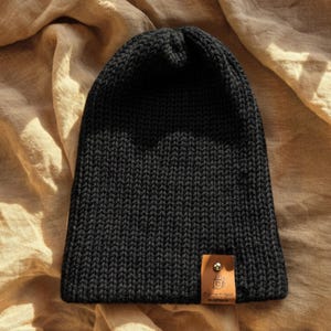 Op de afbeelding: Een effen zwarte gebreide muts met een verticale ribbelstructuur en een kleine, rechthoekige beige leren patch aan de onderrand. De patch toont een klein icoon van een koffiekopje en de tekst "the bean box" in zwarte inkt.