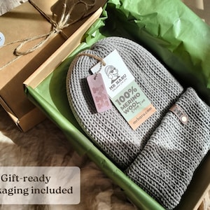 Op de afbeelding: Een grijze gebreide muts en sjaalset in een kartonnen geschenkdoos. De muts heeft een wit label met de tekst MR HARU 100% MERINO WOOL en een klein roze label met de letter M. De doos is bekleed met groen vloeipapier voor een nette presentatie.