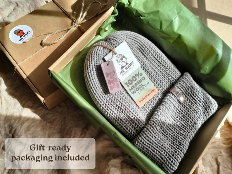 Op de afbeelding: Een grijze gebreide muts en sjaalset in een kartonnen geschenkdoos. De muts heeft een wit label met de tekst MR HARU 100% MERINO WOOL en een klein roze label met de letter M. De doos is bekleed met groen vloeipapier voor een nette presentatie.