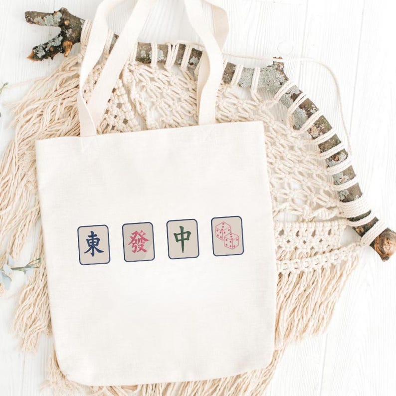 May include: A cream-colored canvas tote bag featuring a graphic print of four rectangular mahjong tiles. The tiles display the characters for East, Green Dragon, and Red Dragon, followed by a tile showing two red dice with black outlines.