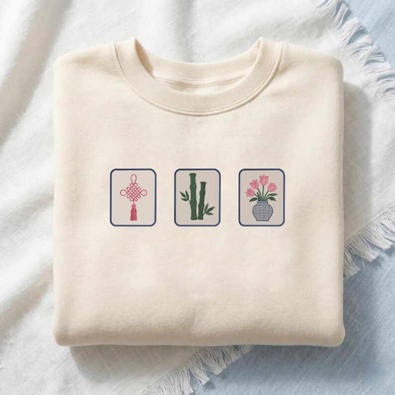 May include: A cream-colored crewneck sweatshirt featuring three embroidered square patches. The patches display a pink Chinese knot, green bamboo stalks, and a vase with pink tulips, each enclosed in a dark blue border on the front chest.