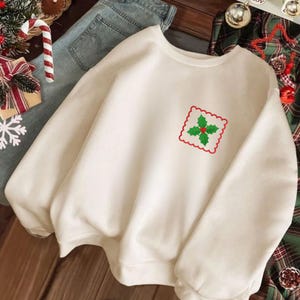 May include: A cream-colored crewneck sweatshirt featuring a small, embroidered square patch on the left chest. The patch has a scalloped red border and displays a green holly leaf design with a single red berry in the center.