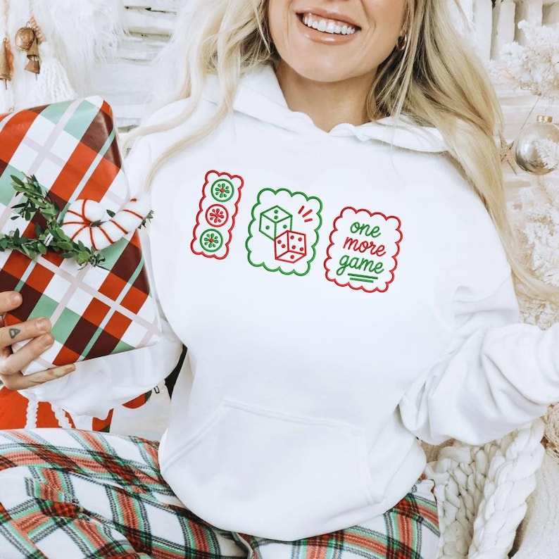 May include: A white hooded sweatshirt featuring a graphic print of three scalloped-edge rectangles. The designs include a vertical strip of three circular ornaments, two dice, and the text "one more game" in red and green ink on the front.