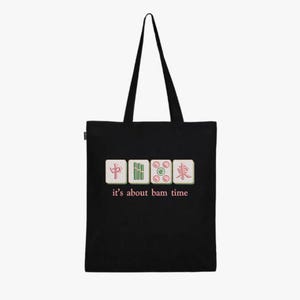 May include: Black tote bag featuring four Mahjong tiles with Chinese characters and the phrase "it's about bam time" in pink. The bag has a simple design with long black handles.