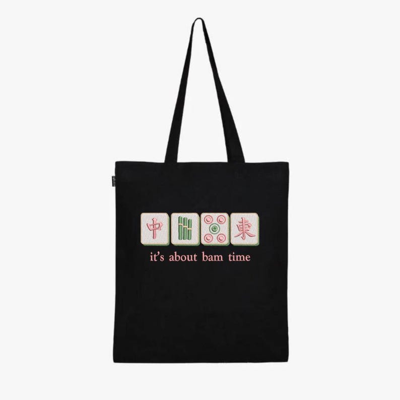 May include: Black tote bag featuring four Mahjong tiles with Chinese characters and the phrase "it's about bam time" in pink. The bag has a simple design with long black handles.