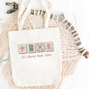 May include: Cream-colored tote bag featuring embroidered Mahjong tiles in red, green, and pink. The phrase "it's about bam time" is embroidered below the tiles in pink. The bag has a simple design with long handles.