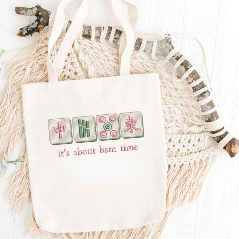 May include: Cream-colored tote bag featuring embroidered Mahjong tiles in red, green, and pink. The phrase "it's about bam time" is embroidered below the tiles in pink. The bag has a simple design with long handles.