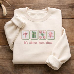 May include: Cream-colored sweatshirt with embroidered Mahjong tiles and the phrase "it's about bam time" in pink thread. The design features four tiles with Chinese characters and bamboo symbols.