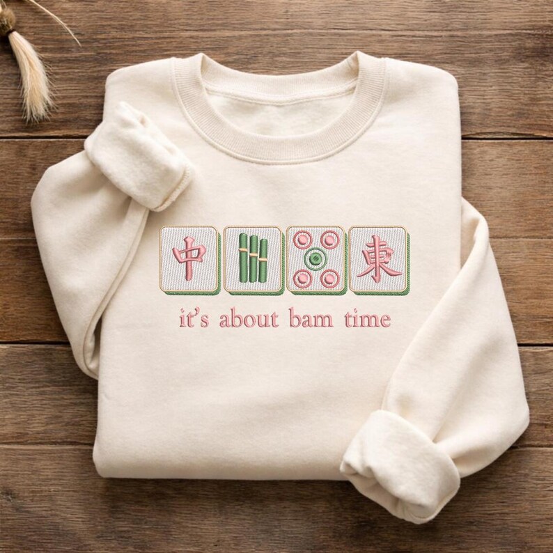 May include: Cream-colored sweatshirt with embroidered Mahjong tiles and the phrase "it's about bam time" in pink thread. The design features four tiles with Chinese characters and bamboo symbols.