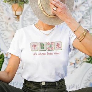 May include: White t-shirt featuring a Mahjong tile design with the text "it's about bam time" in a playful font. The design includes four tiles in green, pink, and red. The shirt is a casual, everyday wear item.