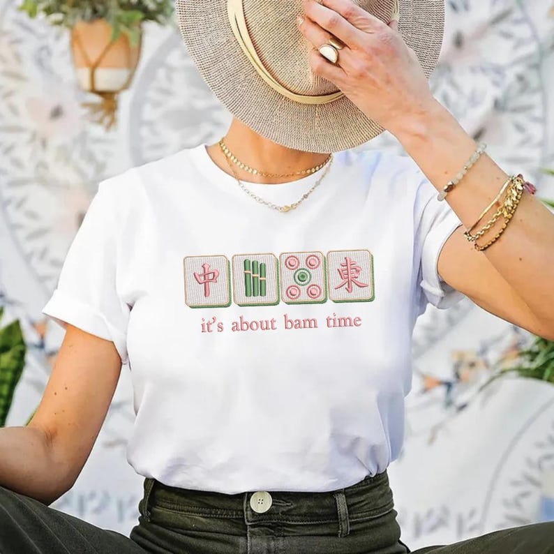 May include: White t-shirt featuring a Mahjong tile design with the text "it's about bam time" in a playful font. The design includes four tiles in green, pink, and red. The shirt is a casual, everyday wear item.