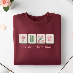 May include: A maroon sweatshirt with embroidered Mahjong tiles and the phrase "it's about bam time." The design features four square tiles with Chinese characters and symbols in green, pink, and white. The sweatshirt is folded neatly on a white surface.
