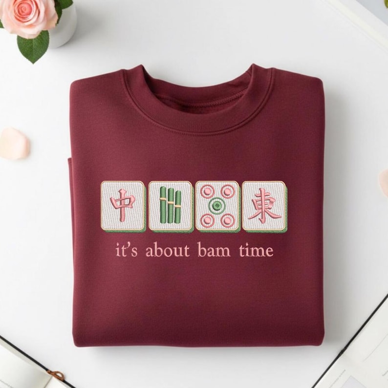 May include: A maroon sweatshirt with embroidered Mahjong tiles and the phrase "it's about bam time." The design features four square tiles with Chinese characters and symbols in green, pink, and white. The sweatshirt is folded neatly on a white surface.