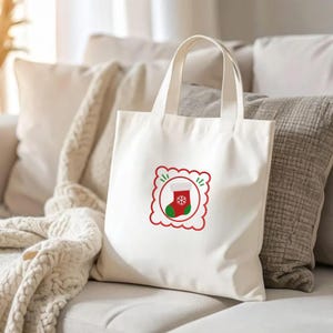 May include: A cream-colored canvas tote bag featuring a centered graphic of a red and green Christmas stocking with a white snowflake design, enclosed within a red scalloped square frame with small green accent marks on the top and sides.