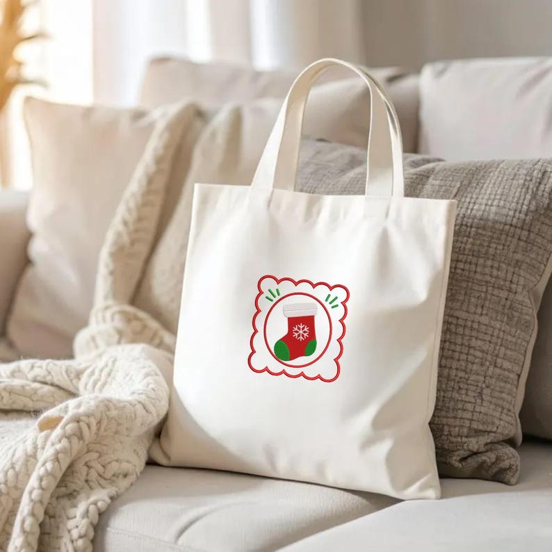 May include: A cream-colored canvas tote bag featuring a centered graphic of a red and green Christmas stocking with a white snowflake design, enclosed within a red scalloped square frame with small green accent marks on the top and sides.