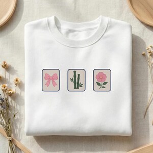 May include: A white crewneck sweatshirt featuring three rectangular embroidered patches. The left patch shows a pink bow, the center patch displays green bamboo stalks, and the right patch depicts a pink flower with green leaves on a light background.