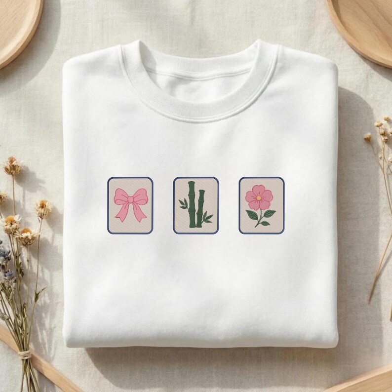 May include: A white crewneck sweatshirt featuring three rectangular embroidered patches. The left patch shows a pink bow, the center patch displays green bamboo stalks, and the right patch depicts a pink flower with green leaves on a light background.
