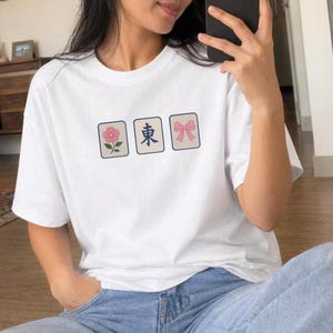 May include: A white short-sleeved t-shirt featuring three rectangular graphics across the chest. The designs include a pink flower, the blue Japanese character for east, and a pink bow, all framed in thin dark borders on the fabric.