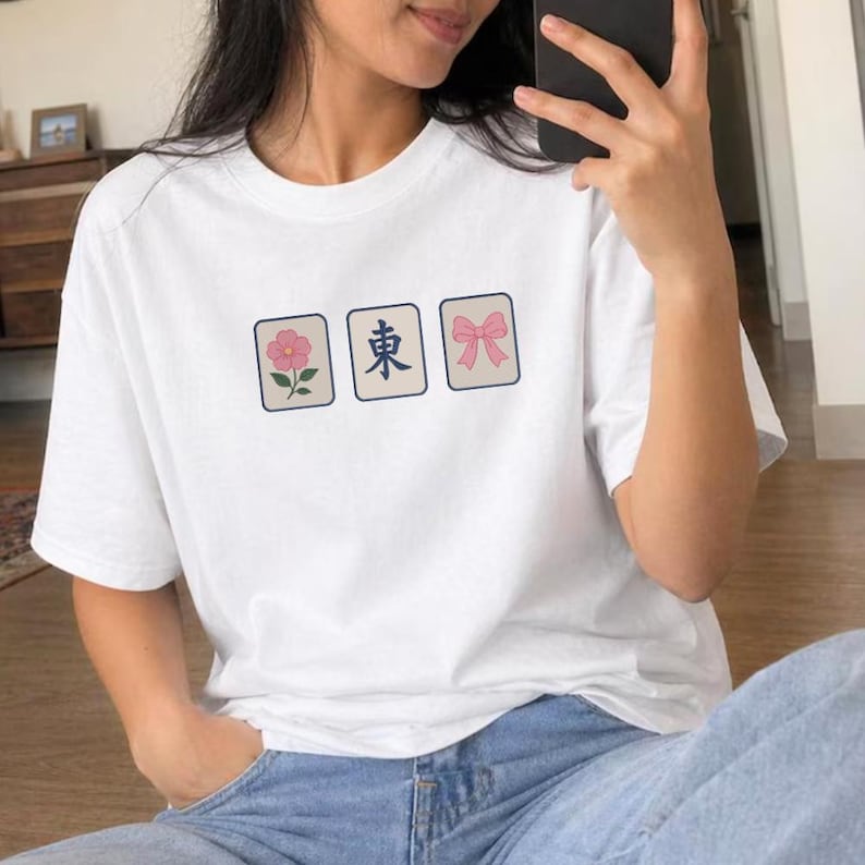 May include: A white short-sleeved t-shirt featuring three rectangular graphics across the chest. The designs include a pink flower, the blue Japanese character for east, and a pink bow, all framed in thin dark borders on the fabric.