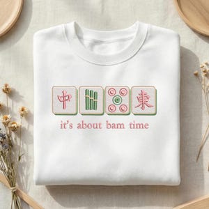May include: White sweatshirt with embroidered Mahjong tiles in green and pink. The text "it's about bam time" is below the tiles. The sweatshirt is a crew neck style.