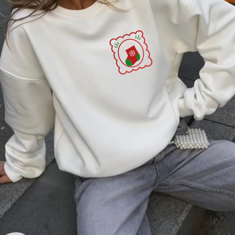 May include: A cream-colored crewneck sweatshirt featuring a small graphic on the upper left chest. The graphic consists of a red scalloped square frame containing a green and red Christmas stocking with a white snowflake detail in the center.
