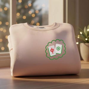 May include: A light pink crewneck sweatshirt featuring an embroidered design on the left chest. The design consists of two mahjong tiles enclosed in a green scalloped border. One tile displays a red Chinese character, and the other a green snowflake.