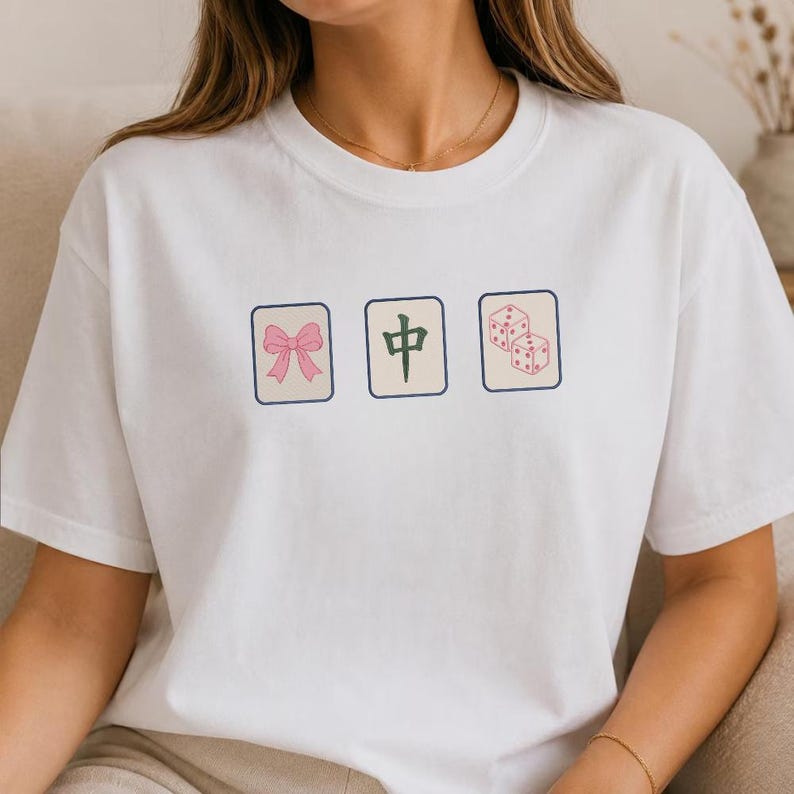 May include: A white short-sleeved t-shirt featuring three rectangular graphics arranged horizontally. The left graphic shows a pink bow, the center graphic displays a green Chinese character, and the right graphic depicts two pink dice.