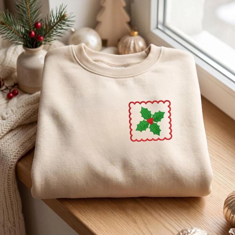 May include: A beige crewneck sweatshirt featuring a small, embroidered square patch on the left chest. The patch has a scalloped red border and contains a detailed green holly leaf design with three small red berries in the center.