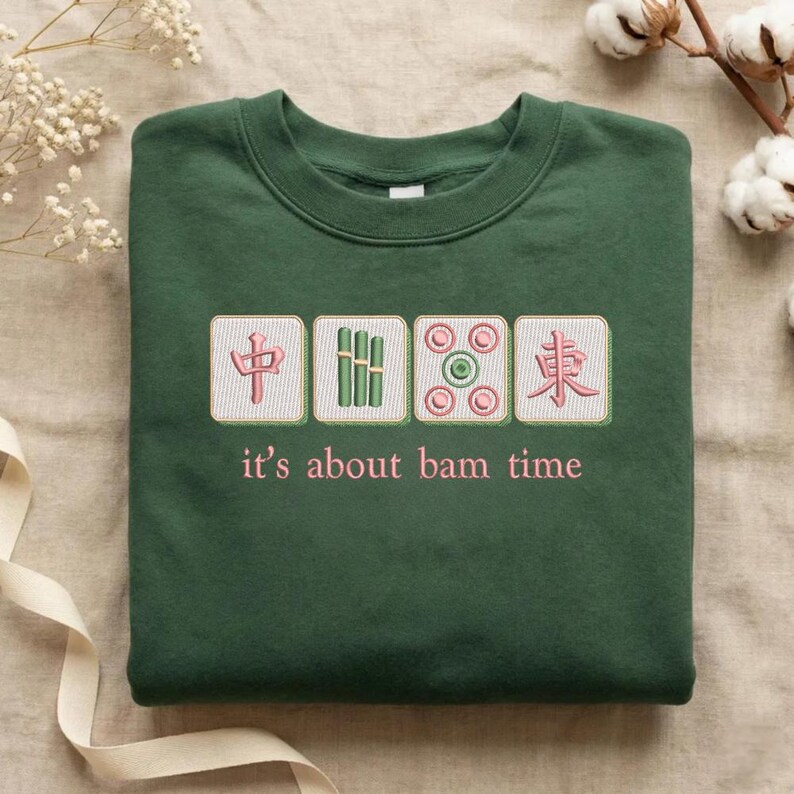 May include: A dark green sweatshirt with embroidered Mahjong tiles and the phrase "it's about bam time" in pink. The design features four tiles with Chinese characters and bamboo symbols.