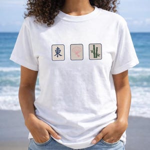May include: A white short-sleeve t-shirt featuring three rectangular graphics across the chest. The designs include a blue East character, a pink folding fan, and green bamboo stalks, all set against a light beige background on the shirt.