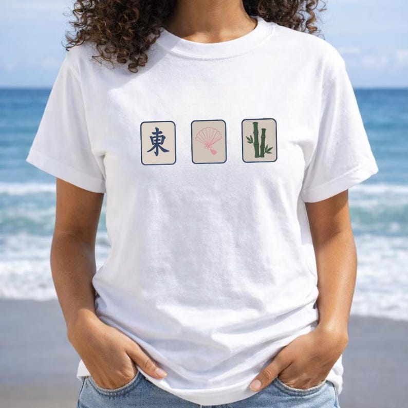 May include: A white short-sleeve t-shirt featuring three rectangular graphics across the chest. The designs include a blue East character, a pink folding fan, and green bamboo stalks, all set against a light beige background on the shirt.