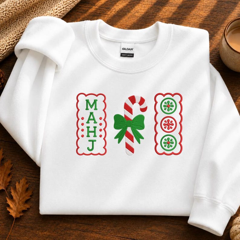 May include: A white crewneck sweatshirt featuring an embroidered design with the vertical text MAHJ in green, a red and white striped candy cane with a green bow, and three circular green and red floral patterns inside a scalloped border.