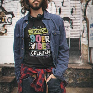 Könnte beinhalten: Schwarzes T-Shirt mit dem Text "VORSICHT 90ER VIBES GELADEN" in farbenfroher Retro-Schrift. Die Person trägt eine blaue Jeansjacke, dunkle Jeans und ein rot-schwarz kariertes Hemd um die Taille gebunden. Kopfhörer liegen um den Hals.