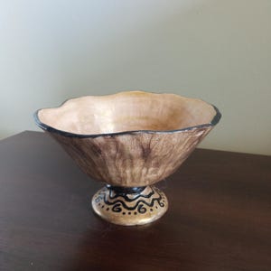 Może przedstawiać: Dekoracyjna miska o teksturowanym, brązowym wyglądzie zewnętrznym i jasnym wnętrzu. Obręcz jest obrysowana na czarno. Misa stoi na podstawie z czarno-złotymi malowidłami. Misa jest prawdopodobnie używana do celów dekoracyjnych.