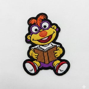 Puede incluir: Parche bordado con un personaje de dibujos animados de piel amarilla, con gafas moradas y una diadema morada. El personaje sonríe y sostiene un libro marrón. El parche tiene un borde negro sobre un fondo blanco.