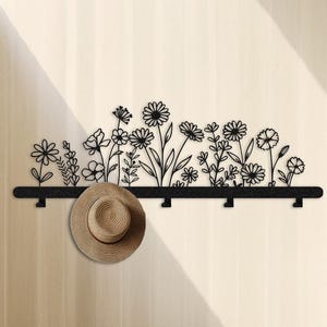May include: Black metal wall-mounted coat rack with a floral design. The rack features a row of hooks and a decorative silhouette of various flowers. A straw hat hangs from one of the hooks, adding a touch of rustic charm to the entryway decor.