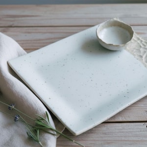 Op de afbeelding: Een vierkante, lichtblauwe keramische serveerschaal met een kleine, ronde kom. De schaal heeft een gespikkelde afwerking en een textuurrand. Een linnen doek en lavendel takjes liggen op de voorgrond. Op een houten oppervlak.