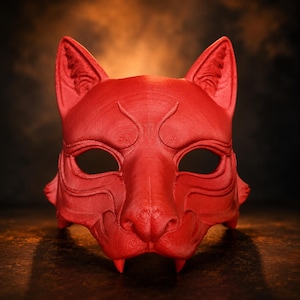 Puede incluir: Una máscara de color rojo brillante con forma de rostro felino, orejas puntiagudas, aberturas para los ojos en forma de almendra y dos pequeños colmillos que sobresalen. La superficie muestra líneas de impresión 3D horizontales.