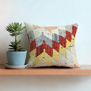 Puede incluir: Cojín decorativo cuadrado con un patrón geométrico en rojo, amarillo, gris y blanco. Se coloca en una estantería de madera junto a una planta suculenta en maceta en una maceta azul claro.