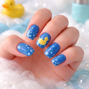 May include: A hand with blue nail polish decorated with white bubbles and a yellow rubber duck design. The nails are surrounded by bubbles and a yellow rubber duck toy.