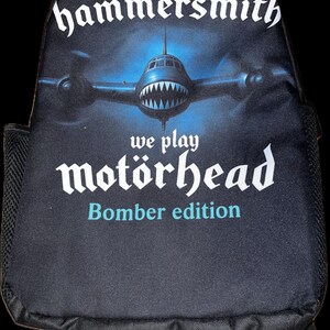 Könnte beinhalten: Schwarzer Rucksack mit einer stilisierten Abbildung eines Bombenflugzeugs mit einem haifischähnlichen Maul. Der Text "hammersmith" steht über dem Flugzeug, mit "we play motörhead" und "Bomber edition" darunter. Der Rucksack hat schwarze Träger und eine Seitentasche aus Mesh.