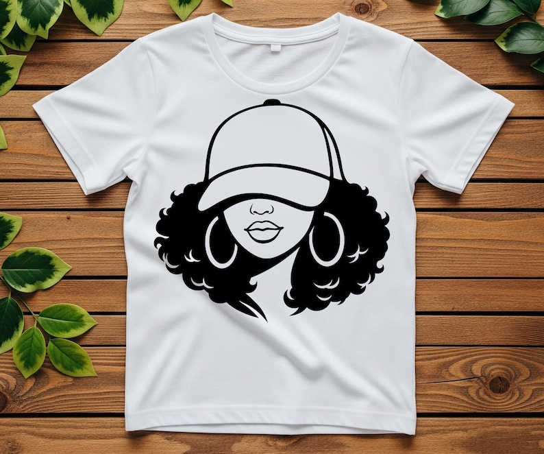 May include: White t-shirt featuring a black and white graphic of a woman wearing a baseball cap and hoop earrings. The design showcases a stylized face with curly hair, a cap, and large hoop earrings. The shirt is made of a soft, comfortable material.