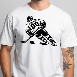 May include: A white short-sleeve t-shirt featuring a black graphic of a hockey player in motion. The player wears a jersey with the text "YOUR NAME" and the number "00" on the back, holding a hockey stick near a puck on the ice surface.