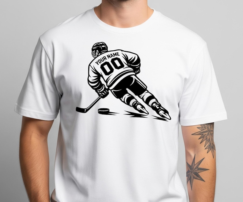 May include: A white short-sleeve t-shirt featuring a black graphic of a hockey player in motion. The player wears a jersey with the text "YOUR NAME" and the number "00" on the back, holding a hockey stick near a puck on the ice surface.