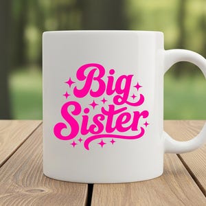 May include: White ceramic mug with a curved handle. The mug features the words "Big Sister" in bright pink, stylized lettering with star accents. The mug is set on a wooden surface with a blurred green background.