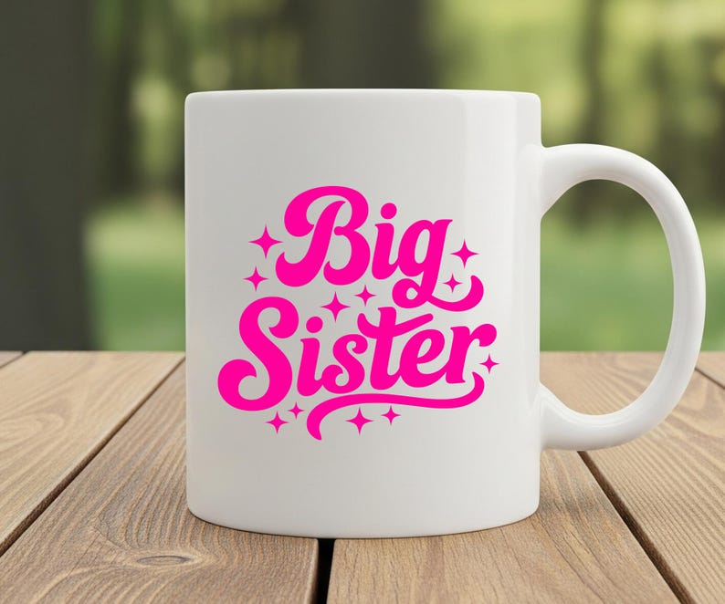 May include: White ceramic mug with a curved handle. The mug features the words "Big Sister" in bright pink, stylized lettering with star accents. The mug is set on a wooden surface with a blurred green background.